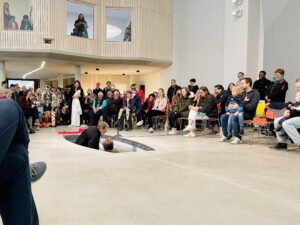 Menschen sitzen im Halbkreis im großen Saal vor Taufbecken, in dem eine Tauf stattfindet