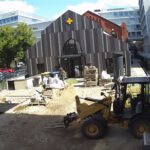 Vor dem Gebäude sind noch von Erde bedeckte Flächen sowie Baufahrzeuge sichtbar.