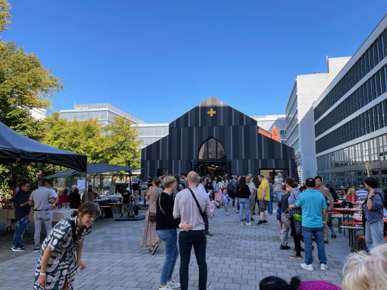 Außenansicht der Baptistenkirche Wedding. Gebäude mit Spitzdach, gelben Kreuz im Giebel und rechteckiger Verkleidung in unterschiedlichen Grautönen. Davor viele Menschen und Essensstände.