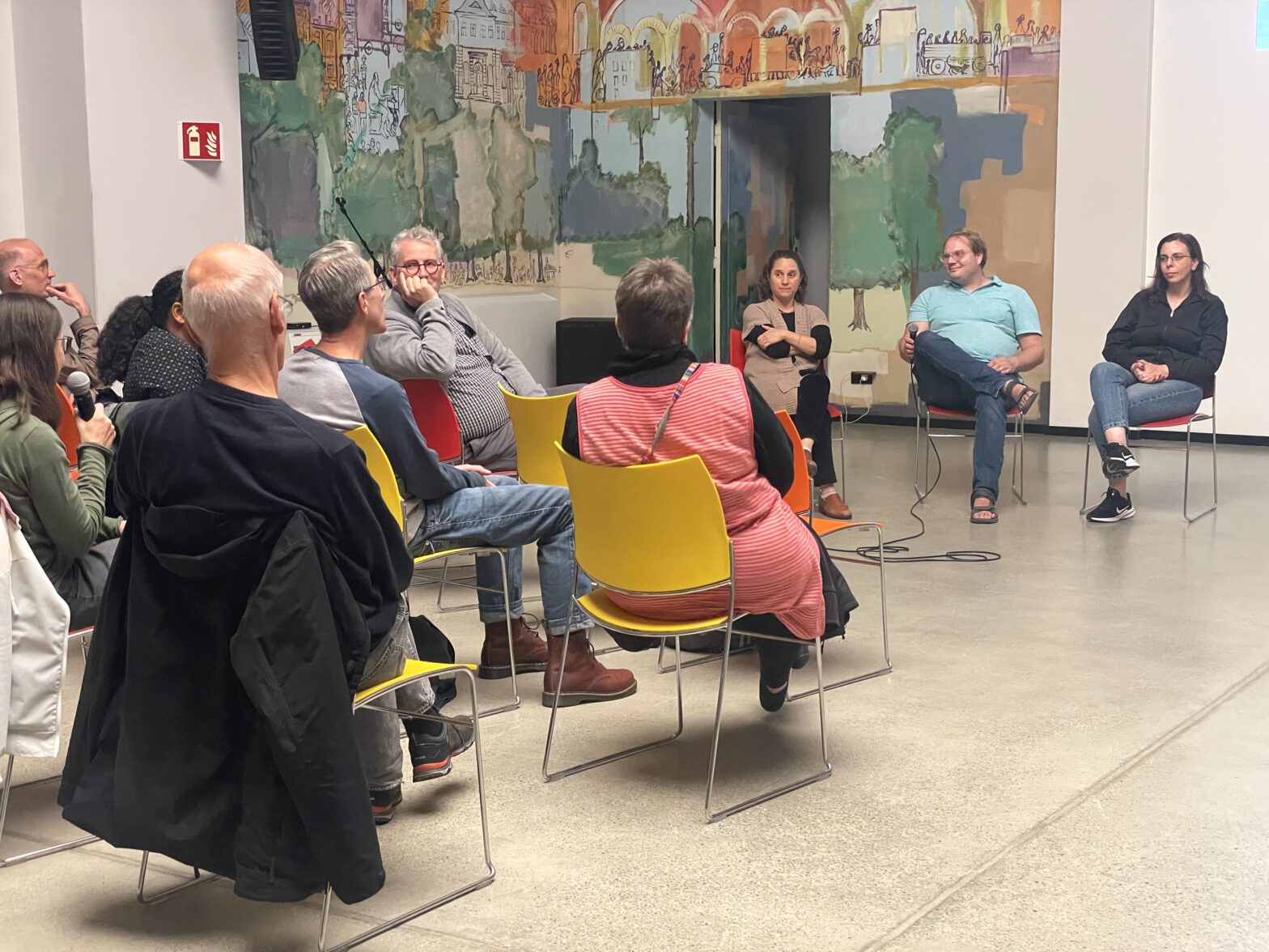 Drei Menschen sitzen vor einem kleinen Pupblikum und sprechen in ein Mikrofon.