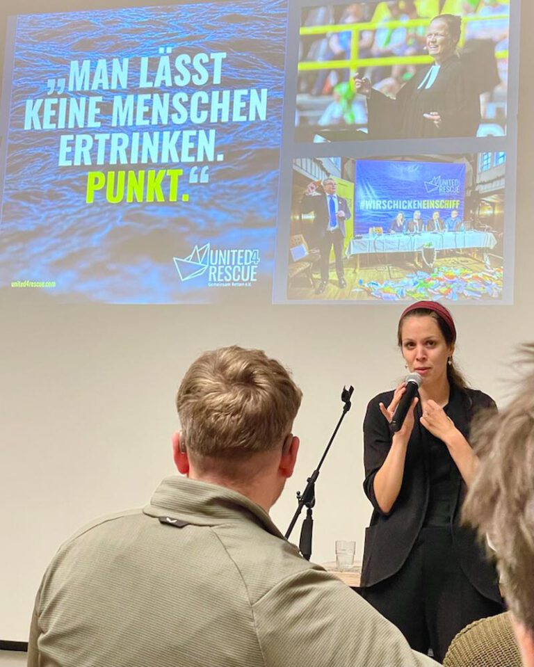 Portrait Annika Schlingheider mit Mikrofon in der Hand vor einem projezierten Bild mit der Aufschrift "Man lässt keine Menschen ertrinken. Punkt."