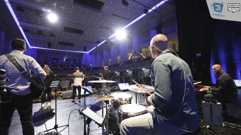 Blick von der Bühne durch Musiker:innen hindurch auf einen großen Saal mit mehreren hundert Menschen.